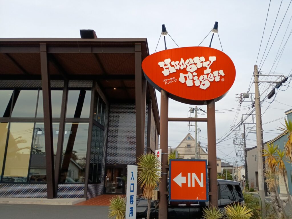 ハングリータイガー湘南辻堂店の店舗外観。曇り空の下、巨大なオレンジ色の楕円形看板に白いロゴと「ステーキとハンバーグ」の文字。大きなガラス窓とレンガ壁の建物、手前に「IN」の矢印看板と「入口専用」の看板