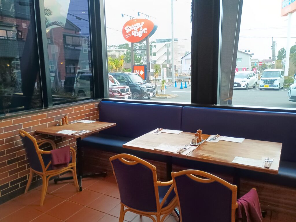 ハングリータイガー湘南辻堂店の店内、窓際の席。テラコッタ色のタイル床、レンガ壁、濃い青紫色のベンチシートと竹編みの椅子。木製テーブルにカトラリーと調味料がセットされ、大きな窓から駐車場とオレンジの看板が見える