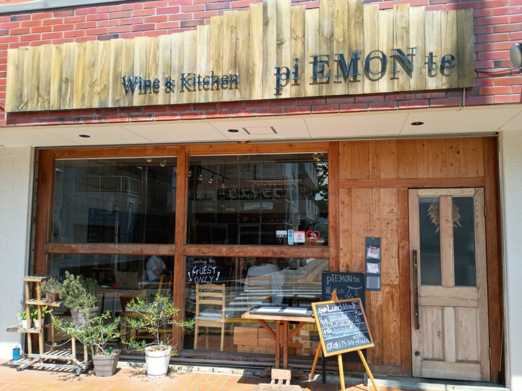 店舗外観:
piEMONteの店舗外観。赤レンガと木材を組み合わせた壁面に「Wine & Kitchen pi EMON te」の看板。正面は大きなガラス張りで、入口前に観葉植物の鉢とランチメニューの黒板が置かれている