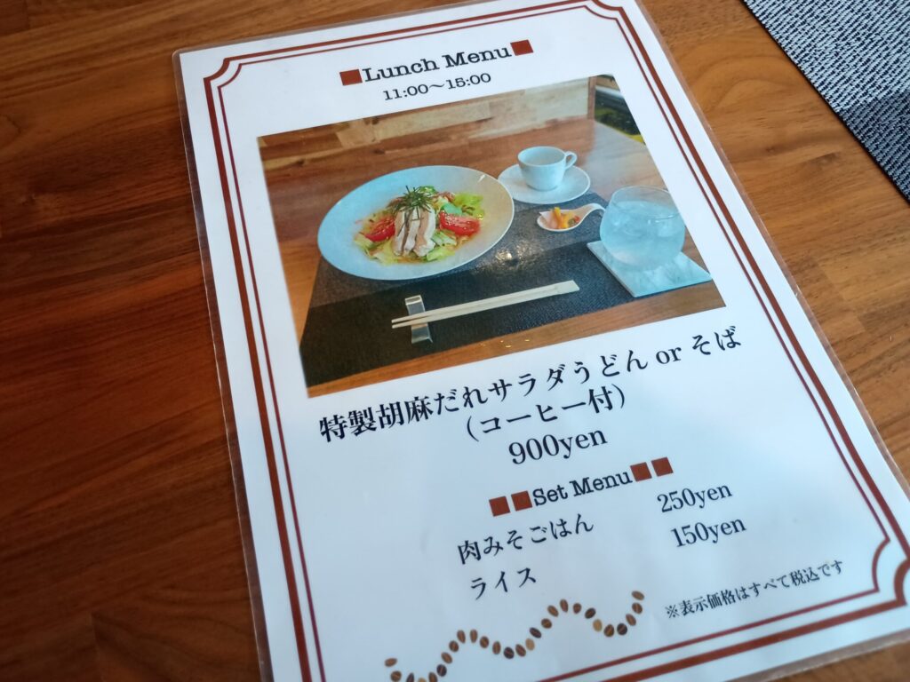 ランチメニュー表。
ラミネート加工されたランチメニュー表。「Lunch Menu 11:00〜15:00」の見出しの下に、特製胡麻だれサラダうどんの写真。「特製胡麻だれサラダうどん or そば（コーヒー付）900yen」、セットメニューとして肉みそごはん250円・ライス150円の記載。「表示価格はすべて税込です」の注記。