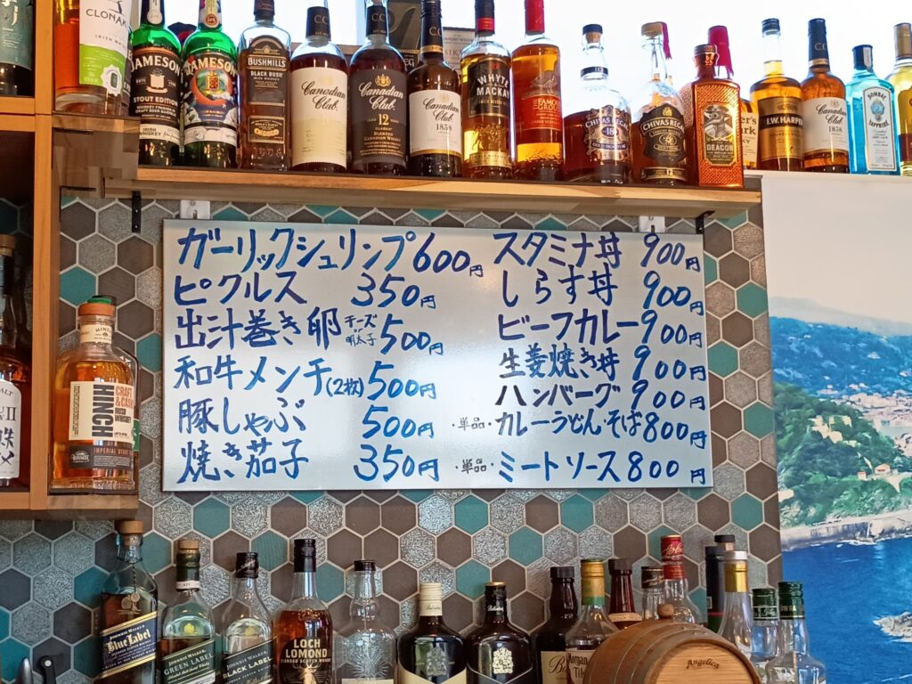 手書きの一品・丼メニュー。
カウンター上の壁に掲げられた手書きのメニューボード。青いマーカーで、ガーリックシュリンプ600円、ピクルス350円、出汁巻き卵500円、和牛メンチ500円などの一品料理と、スタミナ丼・しらす丼・ビーフカレー・生姜焼き丼・ハンバーグが各900円と書かれている。上の棚にはジェムソンやカナディアンクラブなどのウイスキーボトル、右手には湘南の海岸を写したパネル。