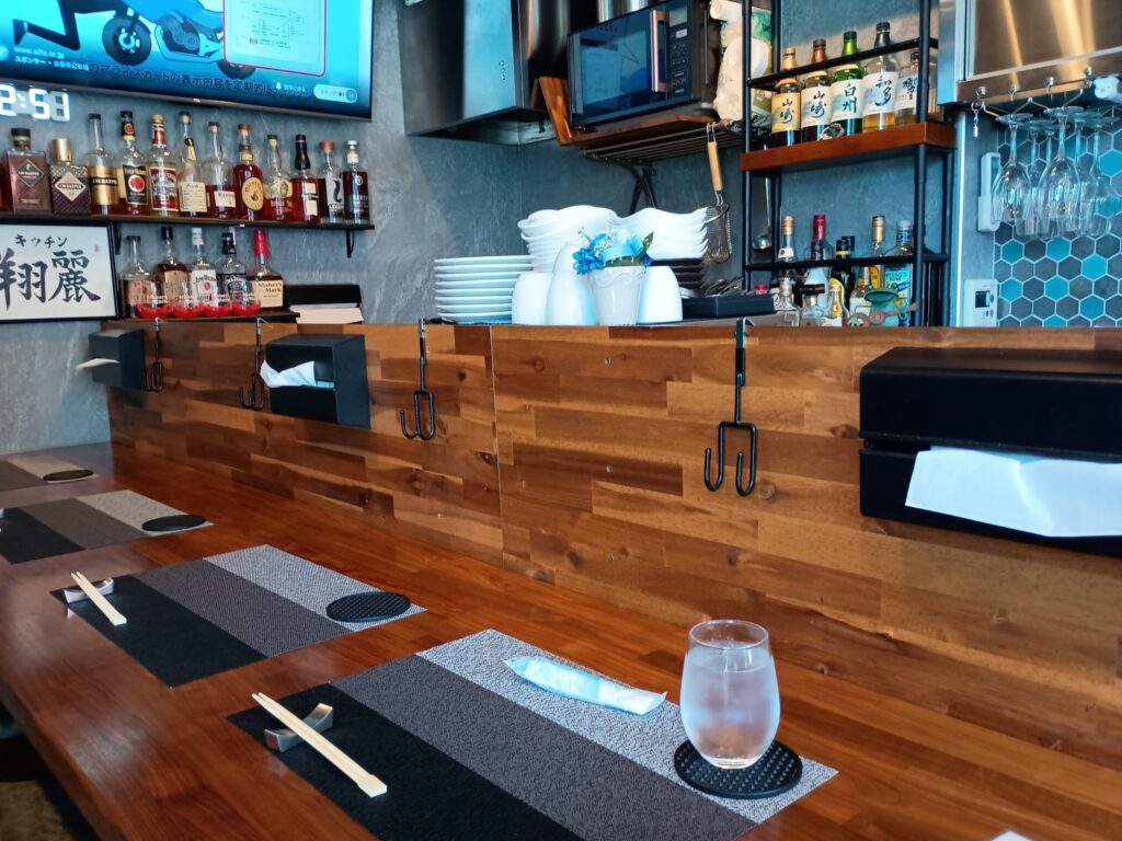 店内・カウンター席。
飴色に磨かれた一枚板の木製カウンター。手前にはランチョンマット、箸置きと割り箸、氷の入った水のグラスがセットされている。奥の棚にはバーボンを中心としたウイスキーボトルがずらりと並び、左端には「キッチン翔麗」の手書き看板。右上には山崎・白州・知多・響などのジャパニーズウイスキー、天井からは脚付きグラスが吊るされた、ダイニングバーらしい店内。