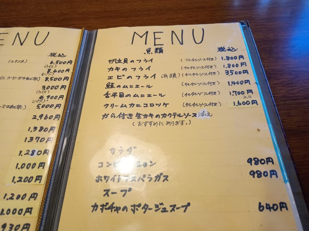 （メニュー）
手書き文字が並ぶ黄色いメニュー表。魚類のページで、帆立貝のフライ、カキのフライ、エビのフライ、鮭のムニエール、舌平目のムニエール、クリームカニコロッケなどが並ぶ。価格は1,400円〜3,500円（税込）。