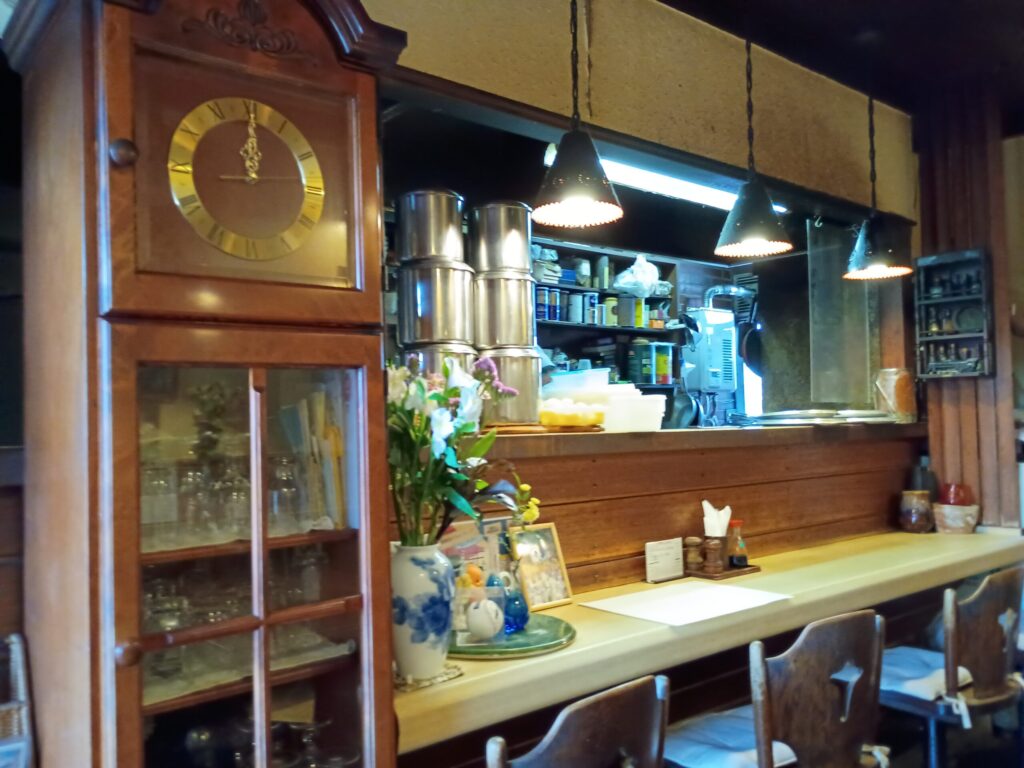 （店内カウンター）
飴色に輝く木製カウンター。左に大きな柱時計。奥の厨房が窓越しに見える。