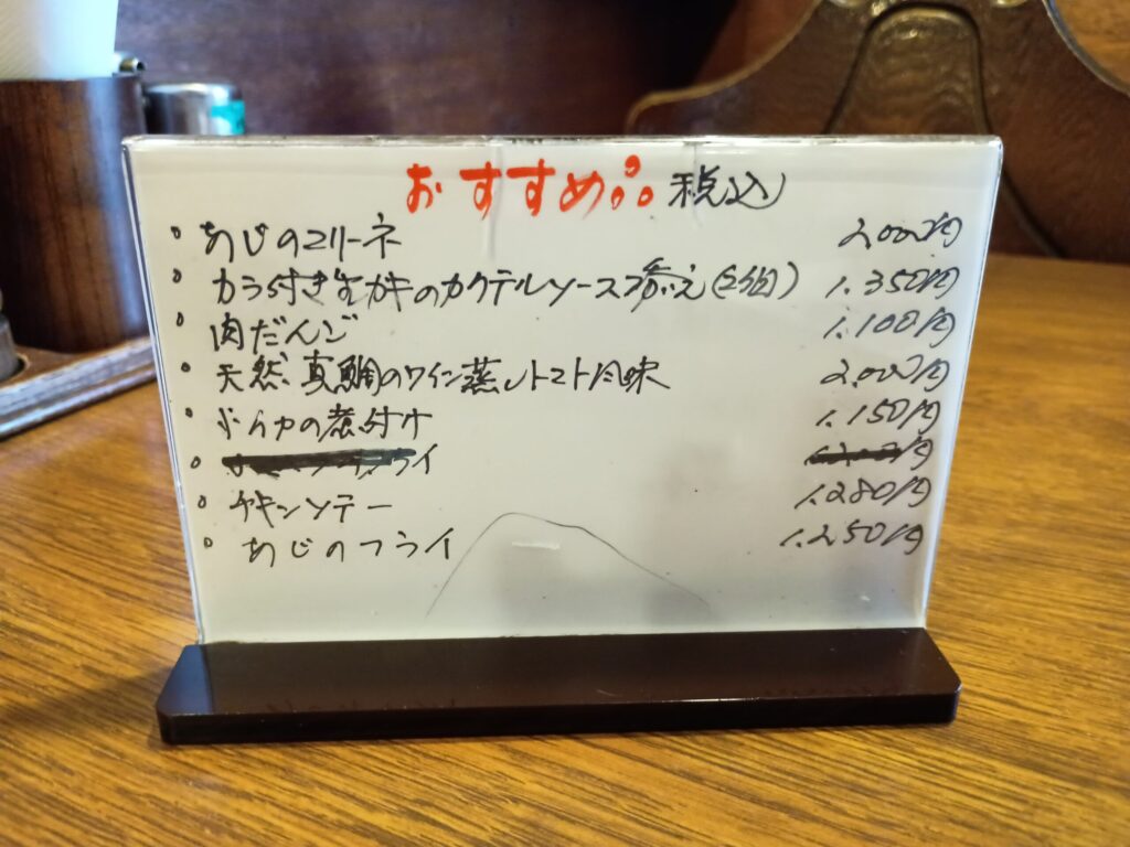（おすすめボード）
テーブルに置かれたアクリル製のおすすめメニュー立て。赤字で「おすすめ。税込」と書かれ、あじのマリーネ、カラ付き生カキのカクテルソース添え、肉だんご、天然真鯛のワイン蒸しトマト風味、あじのフライなどが手書きで記載。