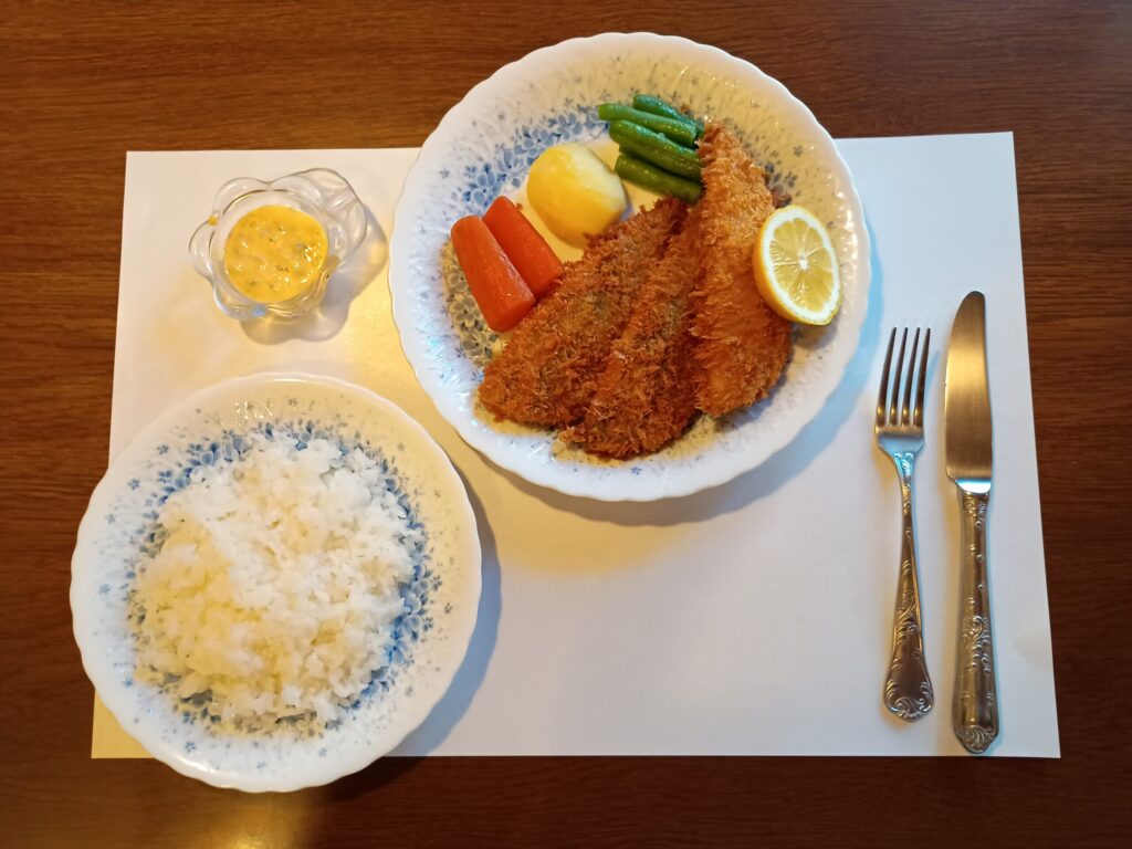 （料理引き）
白いプレースマットの上に、あじのフライの皿とライスの皿を配置した全体図。右にフォークとナイフ。