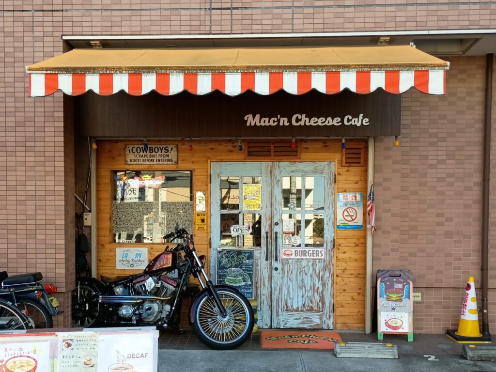 （店舗外観）
マッケンチーズカフェの店舗外観。赤白ストライプの庇、木製のドア、入口前に停まったハーレーダビッドソン。「BURGERS」のサインやアメリカ国旗が飾られたアメリカンな店構え
