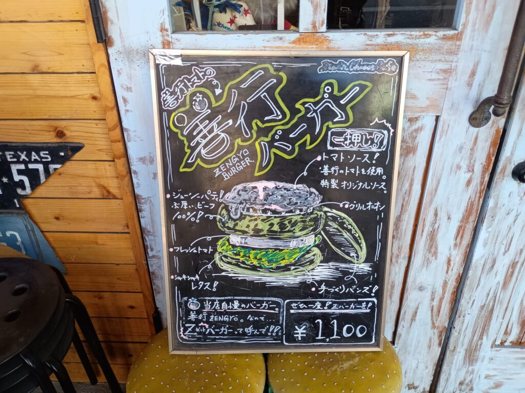 （善行バーガー黒板）
店頭の黒板に描かれた「善行バーガー」のイラスト看板。善行のトマトを使用した特製オリジナルソース、ビーフ100%のジューシーパティ、手づくりバンズなどのアピールポイントと、¥1,100の価格表示