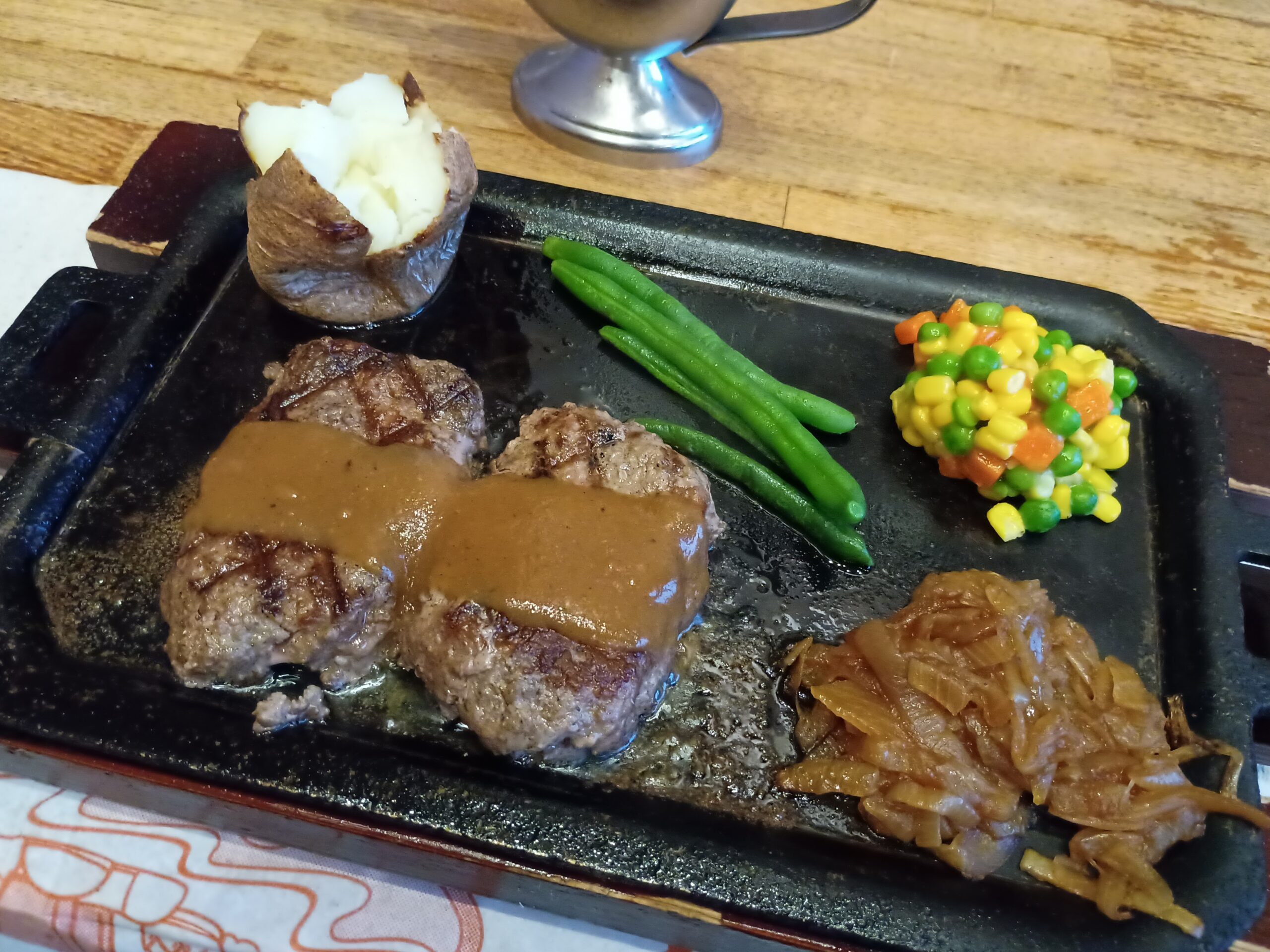 熱々の鉄板にのったハングリータイガーのオリジナルハンバーグステーキ。焦げ目のついた粗挽きハンバーグ2つにブラウンソースがかかり、ベイクドポテト、さやいんげん、ミックスベジタブル、炒めた玉ねぎが添えられている