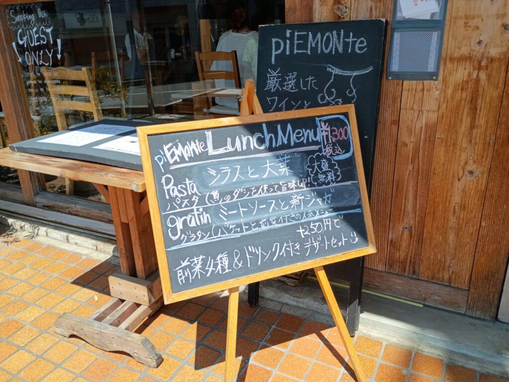 店頭の黒板ランチメニュー:
店頭に置かれたイーゼルの黒板。白チョークで「piEMONte Lunch Menu ¥1,300(税込)」、パスタ「シラスと大葉」、グラタン「ミートソースと新ジャガ」、前菜4種&ドリンク付き、+250円でデザートセットと書かれている
