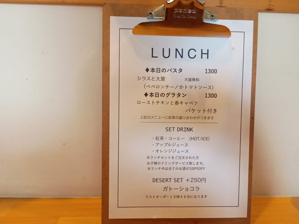 ランチメニュー表:
クリップボードに挟まれたランチメニュー表。本日のパスタ1300円、本日のグラタン1300円、前菜の盛り合わせ付き、SET DRINK(紅茶・コーヒー・ジュース)、DESERT SET +250円 ガトーショコラ、ラストオーダー13時40分