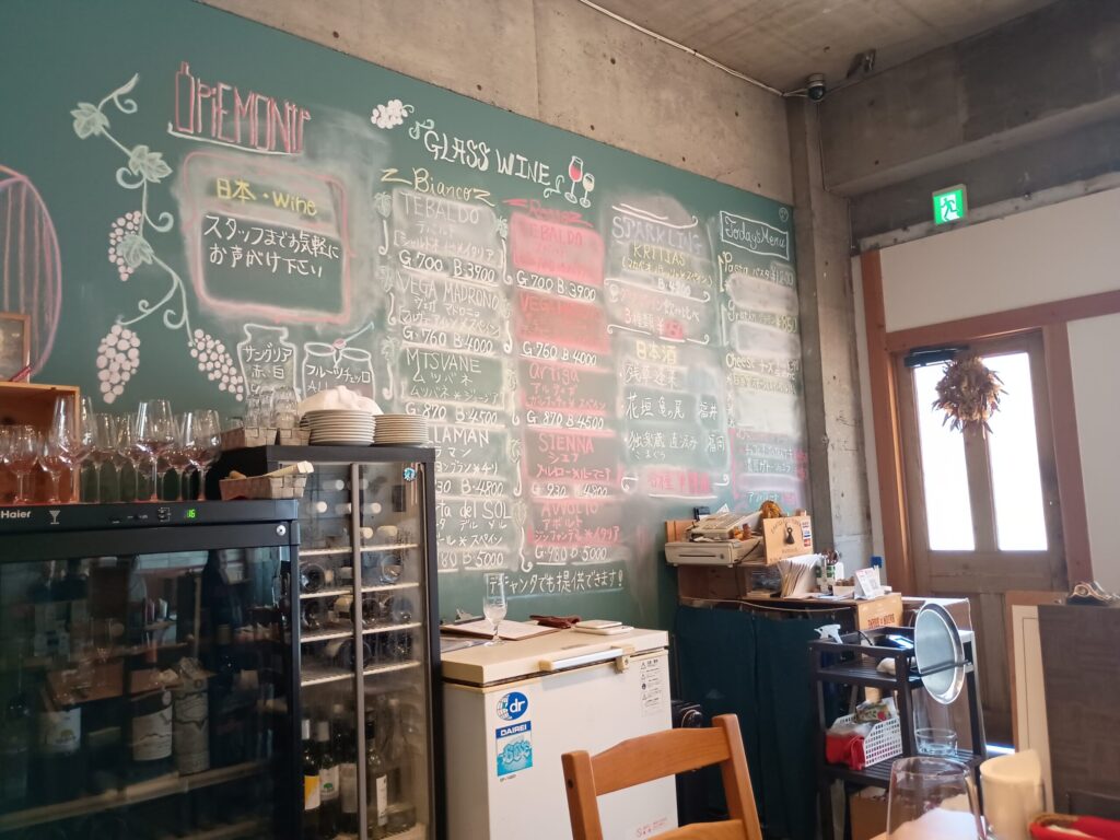 グラスワインの黒板:
店内の緑色の壁一面に手描きされたグラスワインのリスト。ビアンコ(白)、ロッソ(赤)、スパークリング、日本酒の銘柄と価格がチョークで書かれ、ブドウやワインボトルのイラストも添えられている。手前にワインセラーとグラスが並ぶ