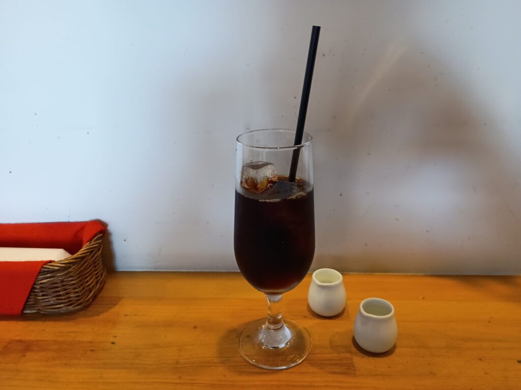 アイスコーヒー:
脚付きグラスに入ったアイスコーヒー。黒いストローが添えられ、横に白い陶器のミルクピッチャーとガムシロップ入れが2つ並んでいる